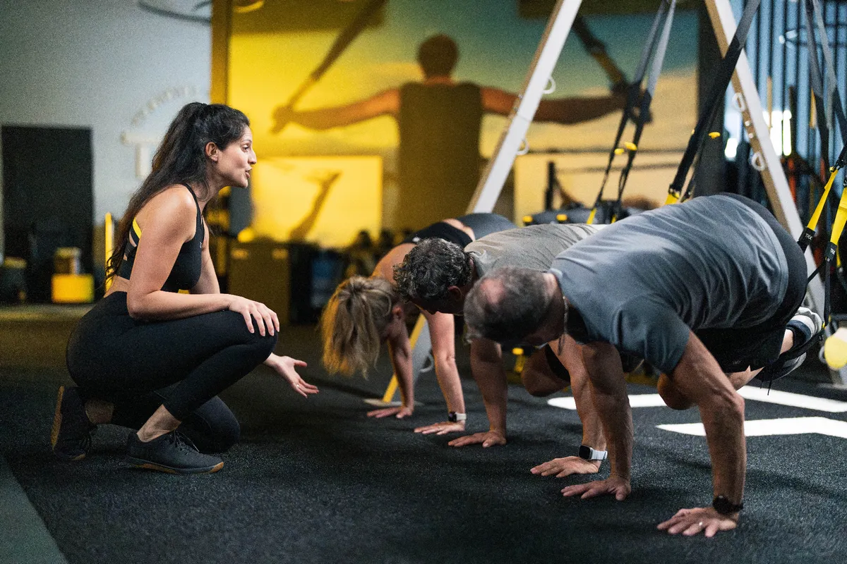 A coach kneeling to cue members holding a TRX plank